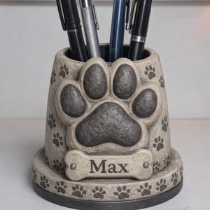 Puede incluir: Un portalápices beige con un diseño de gran huella de pata y huellas de pata más pequeñas alrededor. El nombre "Max" se muestra en una placa con forma de hueso. Varios bolígrafos en el interior.