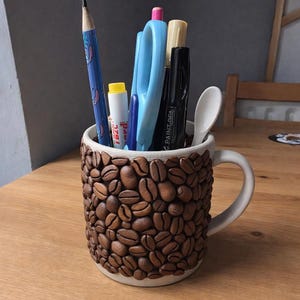 Puede incluir: Una taza de cerámica blanca decorada con granos de café esculpidos. La taza contiene varios artículos de papelería, como un lápiz azul, tijeras y bolígrafos. La taza está sobre una superficie de madera.
