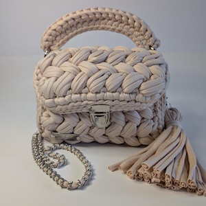 May include: A beige crochet handbag with a braided design, a silver clasp, and a matching handle. The bag includes a silver chain strap and a beige tassel. The bag is approximately 8 inches wide and 6 inches tall.