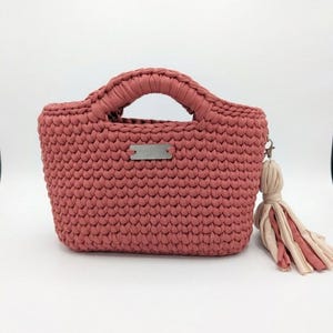 May include: A dusty rose crochet handbag with a rounded handle and a small rectangular tag. A tassel with pink and beige fabric hangs from the side. The bag is a small tote style.