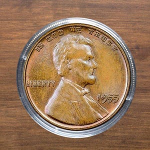 Puede incluir: Una moneda de centavo Lincoln de 1955 de color cobre, dentro de un soporte de plástico transparente. La moneda presenta un perfil de un hombre y las palabras "LIBERTY" e "IN GOD WE TRUST". El año "1955" es visible.