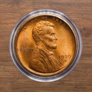 Puede incluir: Una moneda de cobre Lincoln cent de 1909 S, dentro de un soporte protector transparente. La moneda presenta un perfil de Abraham Lincoln, con las palabras "IN GOD WE TRUST" y "LIBERTY". El fondo es de madera.