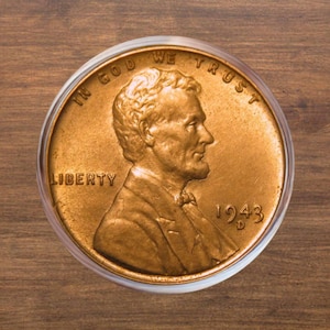 Puede incluir: Una moneda de un centavo Lincoln de 1943 de color cobre, dentro de un soporte de plástico transparente. La moneda presenta el perfil de Abraham Lincoln, las palabras "IN GOD WE TRUST" y "LIBERTY", y la fecha "1943".