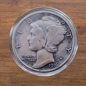 May include: A silver coin encased in a clear plastic holder. The coin features a profile of a woman with winged hair, the words "LIBERTY" and "IN GOD WE TRUST", and the year "1916". The coin is set against a wooden background.