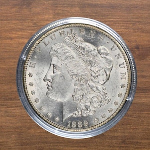 Puede incluir: Una moneda de plata Morgan de 1889, dentro de un protector. La moneda muestra el perfil de una mujer y las inscripciones "E PLURIBUS UNUM" y "LIBERTY". Sobre un fondo de madera.