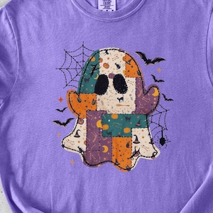 May include: A violet short-sleeve t-shirt featuring a central graphic of a ghost composed of various patterned patches in orange, teal, and cream. The ghost design includes black eyes, spiderwebs, bats, and small pumpkins on a violet fabric background.