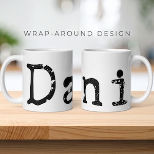 May include: Two white ceramic mugs featuring a wrap-around design with the name Dani printed in a large, black, distressed, textured font. The letters have a weathered, worn appearance, and the mugs are displayed side-by-side on a wooden surface.