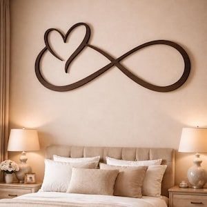 Può includere: Un'opera d'arte da parete in legno marrone scuro con un cuore intrecciato a un simbolo dell'infinito, montata sopra un letto. La camera da letto ha una palette di colori neutri con biancheria da letto beige, cuscini e due lampade da comodino.