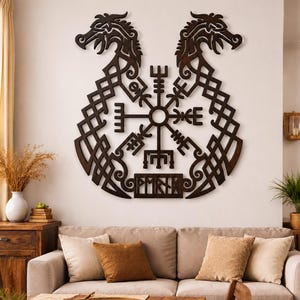 Peut inclure: Une décoration murale en bois brun foncé représentant deux têtes de dragon symétriques tournées vers l'extérieur, encadrant un symbole de boussole Vegvisir central. Sous la boussole, une plaque rectangulaire affiche les runes PMRTM découpées.