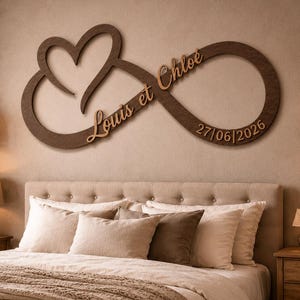 Può includere: Una decorazione da parete in legno marrone scuro a forma di simbolo dell'infinito combinato con un cuore. Il pezzo presenta il testo "Louis et Chloe" in carattere corsivo e la data "27/06/2026" in carattere sans-serif, entrambi in legno chiaro.