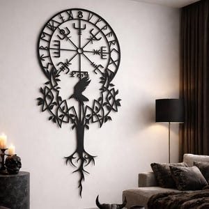 Op de afbeelding: Zwart metalen wandkunst met een Viking kompas ontwerp met runen, een raaf en een boommotief. Het kunstwerk is bevestigd aan een witte muur in een woonkamer, wat een vleugje Scandinavische stijl aan het decor toevoegt.