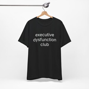 May include: Black t-shirt with the white text "executive dysfunction club". The shirt is hanging on a wooden hanger against a white background. The t-shirt is a crew neck style.