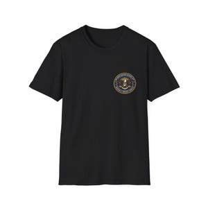 May include: A black short-sleeved t-shirt featuring a circular emblem on the left chest. The emblem displays a central eagle graphic with text reading Non-Human Biologics Recovery Unit and Department of Aerospace Anomalies in a gold and blue design.