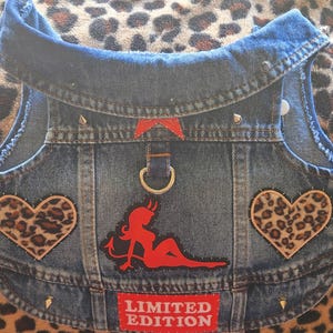 May include: A blue denim dog vest with a decorative design. The vest features two leopard print heart patches, a red devil woman patch, and a red "LIMITED EDITION" patch. The vest has silver metal studs and a metal ring.