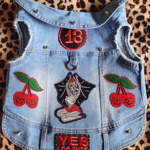 May include: Light blue denim dog vest with patches and metal studs. Patches include a red "13" circle, two cherry skull designs, and a bat-winged figure. The text "YES I DO BITE" is also featured.