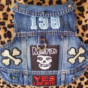 Könnte beinhalten: Eine blaue Hunde-Jeansweste mit der Zahl "138" in Hellblau. Die Weste hat Aufnäher mit einem Totenkopf, gekreuzten Knochen und dem Text "YES I DO BITE". Die Weste hat einen Metallring und dekorative Nieten.