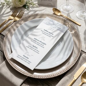 Pode incluir: Um cartão de menu retangular branco com texto em azul escuro intitulado The Menu, listando Buffalo caprese, salmone alla griglia, tagliata e tiramisu, colocado num prato de jantar branco com borda de contas, rodeado por talheres dourados.