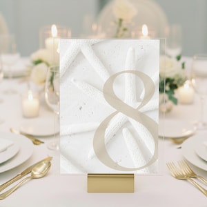 May include: A clear acrylic table number sign featuring a white starfish pattern background and a large, semi-transparent beige number eight printed in the center, supported by a rectangular gold-toned metal base stand.