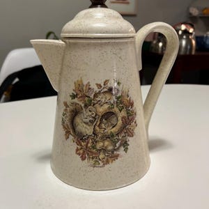 以下が含まれることがあります： クリーム色のエナメルコーヒーポット。斑点模様で、リスと秋の葉のデザインが施されています。ポットには茶色の蓋とハンドル、注ぎ口が付いています。「Squirrels」の文字が印刷されています。