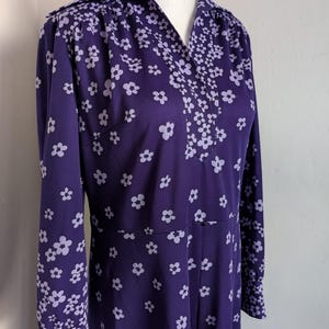 May include: A vintage purple dress with a white floral pattern. The dress features a collared neckline, long sleeves, and a fitted waist. The floral design is concentrated on the collar, sleeves, and lower portion of the dress.
