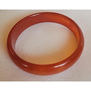 May include: A polished, translucent orange-red bangle bracelet. The smooth, circular bracelet is made of a solid material and has a glossy finish. The bracelet is a simple, elegant piece of jewellery.