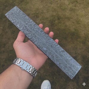 Puede incluir: Una hoja rectangular de acero gris con un patrón arremolinado único. La hoja se sostiene en una mano, mostrando el intrincado diseño. El acero tiene una base gris oscuro con patrones claros y fluidos. La hoja mide aproximadamente 30 cm de largo.