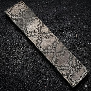 May include: A rectangular metal object with a detailed floral pattern in shades of gray and silver. The design features stylized flower-like shapes and intricate lines. The object is set against a dark, textured background.