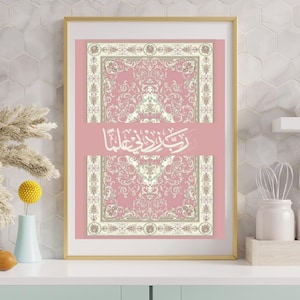 May include: A rectangular wall art print in a gold frame featuring a dusty pink background with two symmetrical, ornate, cream-colored floral patterns. Centered between the patterns is white Arabic calligraphy that reads Rabbi Zidni Ilma.