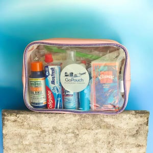 May include: A clear travel pouch filled with toiletries, including toothpaste, mouthwash, tissues, and spray bottles. The pouch is pink with a clear front, and the text "GoPouch" is visible on a white sticker.