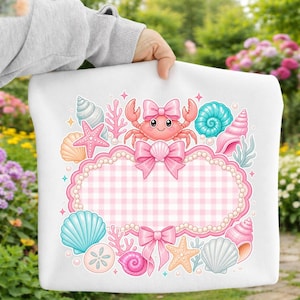 May include: A white sweatshirt featuring a central pink gingham frame decorated with pearls, bows, and a cartoon crab. The design includes various seashells, starfish, and coral in shades of blue, pink, and orange, arranged around the central border.