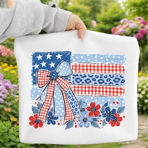 May include: White tote bag featuring a patriotic American flag design. The flag incorporates stars, stripes, and a bow with polka dots and gingham patterns. Red, white, and blue floral accents complete the design. The bag is held by a person.