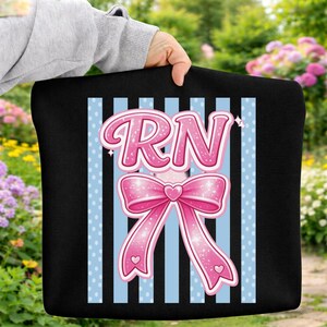 Peut inclure: Sac fourre-tout noir avec un motif comportant les lettres "RN" en rose, au-dessus d'un nœud rose avec un cœur au centre. Le fond présente des rayures verticales bleu clair avec des pois blancs. Le sac est tenu par une personne.