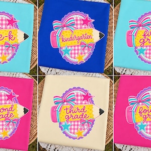 Könnte beinhalten: Eine Sammlung von sechs T-Shirts in verschiedenen Farben, die jeweils eine Grafik eines gelben Bleistifts, ein rosa-weiß kariertes Oval, Schleifen und Sterne zeigen. Die T-Shirts tragen die Aufschriften pre-k, kindergarten, first grade, second grade, third grade und fourth grade.