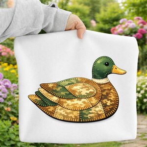 May include: A white tote bag featuring a detailed illustration of a duck. The duck has a green head, a yellow beak, and a body composed of camouflage-patterned patches in shades of green, brown, and tan. The bag is held by a person in front of a garden.