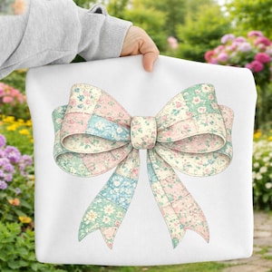 Puede incluir: Un cojín blanco con un diseño de lazo grande. El lazo está hecho de tela de retazos en colores pastel, incluyendo rosa, azul y verde, con patrones florales. El lazo es el foco central de la imagen.