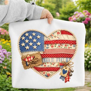 Op de afbeelding: Een wit kussen met een hartvormig ontwerp met een patchwork Amerikaanse vlag. De vlag bevat blauwe stof met witte sterren, rood-witte strepen en bloempatronen. Een klein label met een blauw hart en belletjes hangt aan de onderkant.