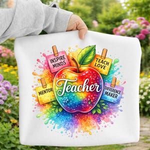 May include: White tote bag featuring a colorful apple design with the word "Teacher" in script. The apple is surrounded by splashes of rainbow colors and small notes that say "Inspire Minds", "Teach Love", "Mentor", and "Difference Maker".