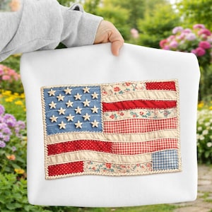 Può includere: Un cuscino bianco con un design a bandiera americana trapuntata. La bandiera ha un campo blu denim con stelle bianche, strisce rosse e floreali e un bordo simile a una corda. Il cuscino è tenuto.