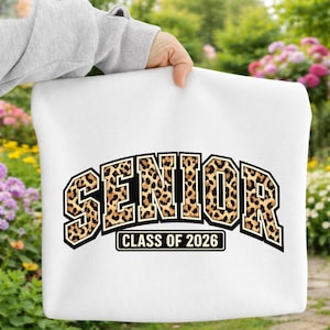 Può includere: Borsa tote bianca con la scritta "SENIOR" in un motivo maculato, bordata di nero. Sotto la parola "SENIOR" c'è la scritta "CLASS OF 2026" in un rettangolo nero. La borsa è tenuta da una persona.