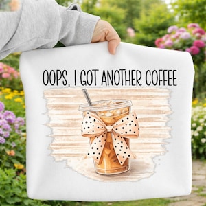 Peut inclure: Coussin blanc avec le texte "OOPS, I GOT ANOTHER COFFEE" en noir. Le coussin présente une illustration d'un café glacé avec un nœud et une paille. Le nœud est beige avec des pois noirs.