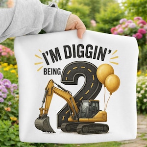 Può includere: Cuscino bianco con una scavatrice gialla e la scritta "I'M DIGGIN' BEING 2". Il numero 2 è disegnato come una strada con palloncini gialli.