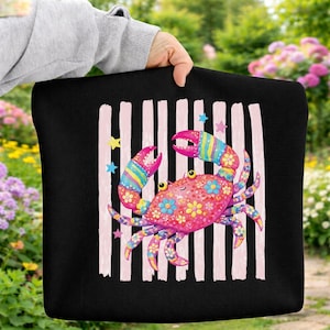May include: Black tote bag featuring a colorful crab design. The crab is pink with floral patterns and rainbow-striped claws. Pink vertical stripes and star accents are on the black background. The bag is held by a person.