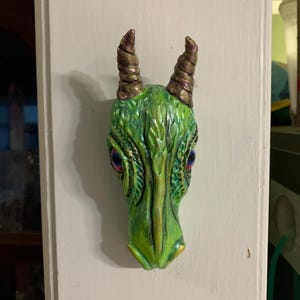 Pode incluir: Uma escultura de cabeça de dragão verde com escamas detalhadas e características texturizadas. O dragão tem chifres castanhos e olhos azuis e roxos. A escultura é montada em uma superfície branca.