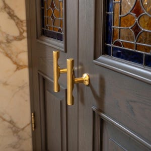 May include: Close-up of a gray wooden door with a gold-colored handle. The door features a stained glass window with shades of blue, orange, and brown. The handle is a horizontal bar with a vertical bar attached.