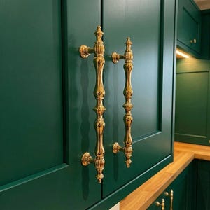 Może przedstawiać: Zbliżenie dwóch ozdobnych, złotych uchwytów do szafek na tle ciemnozielonych drzwi szafki. Uchwyty mają pionową, wydłużoną konstrukcję z dekoracyjnymi detalami. Szafka jest częścią ustawienia kuchennego.