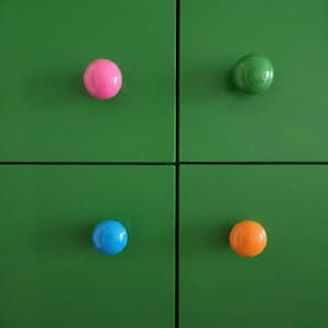 May include: Close-up of a green cabinet with four colorful, round knobs. The knobs are pink, green, blue, and orange. The cabinet doors are a solid, vibrant green color, creating a striking contrast with the knobs.
