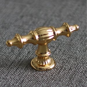 Könnte beinhalten: Ein goldfarbener Schrankgriff mit dekorativem Design. Der Griff hat einen gerippten Mittelteil und verzierte Enden. Der Sockel ist rund und detailliert. Die Hardware ist wahrscheinlich aus Metall und für Möbel konzipiert. Die Oberfläche ist glänzend und reflektierend.