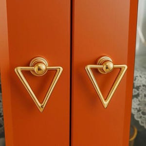 Op de afbeelding: Close-up van twee kastdeuren met bijpassende goudkleurige driehoekige handgrepen. De handgrepen zijn met een cirkelvormige basis en een kleine bol aan de deuren bevestigd. De kastdeuren zijn effen oranje.
