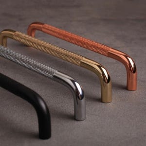 May include: A collection of four cabinet handles in various metallic finishes. The handles are curved with a textured grip and come in black, silver, gold, and copper. The handles are designed for furniture and home decor.