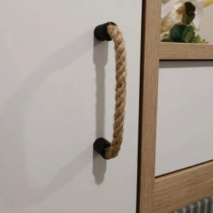 May include: A cabinet pull featuring a natural rope handle. The rope is attached to the cabinet with two black cylindrical mounts. The cabinet is white, and the pull is mounted vertically.